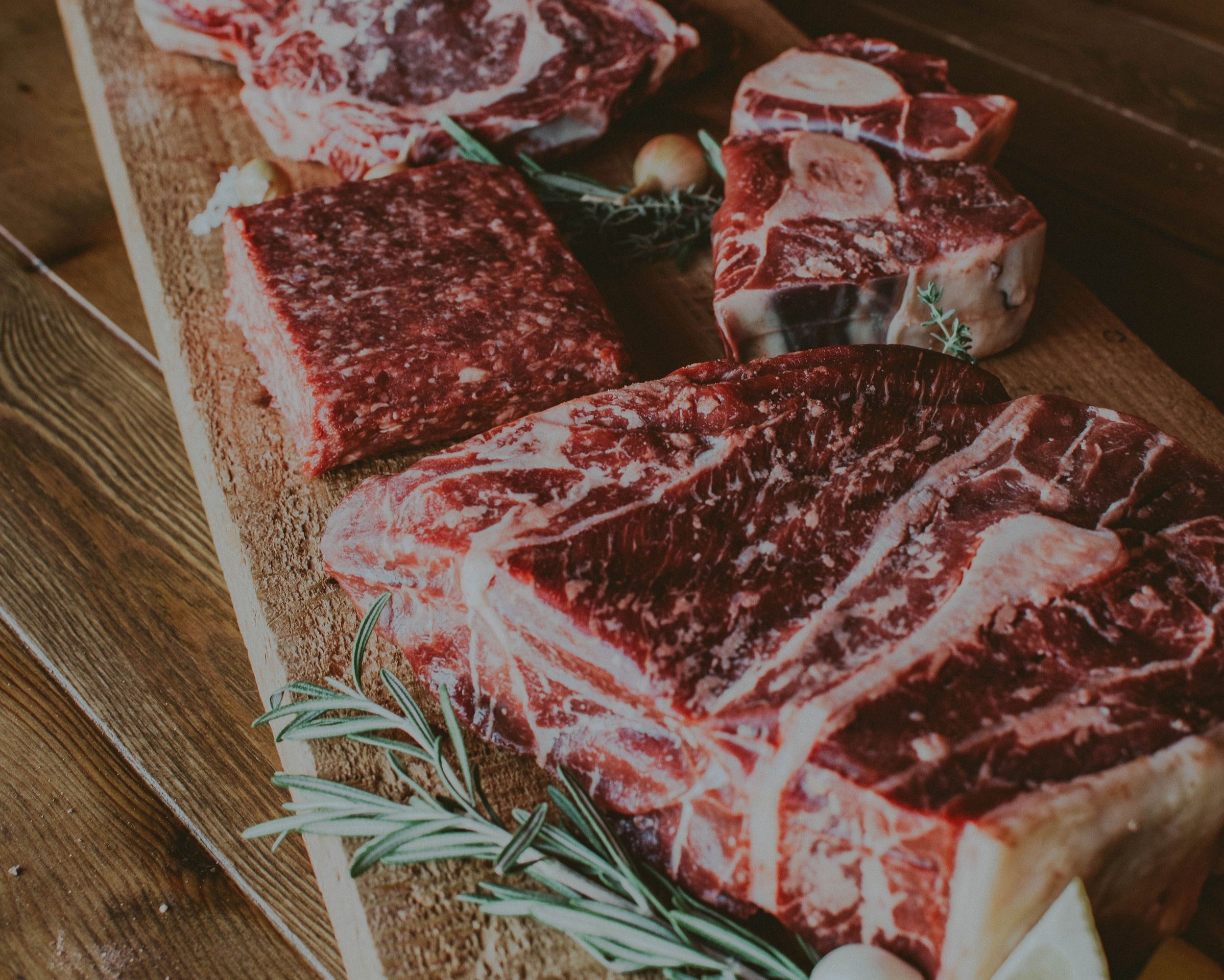 Assorted cuts of fresh, high-quality beef displayed on a wooden cutting board, including ribeye, chuck, and ground beef, garnished with fresh rosemary and garlic cloves.