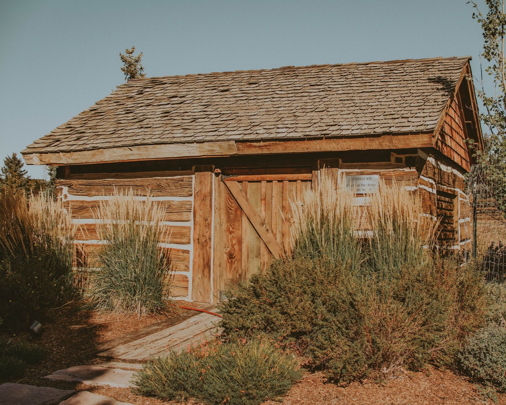 Historic wooden cabin surrounded by lush greenery and ornamental grasses, showcasing rustic architecture and natural landscaping. Ideal for nature lovers and history enthusiasts.