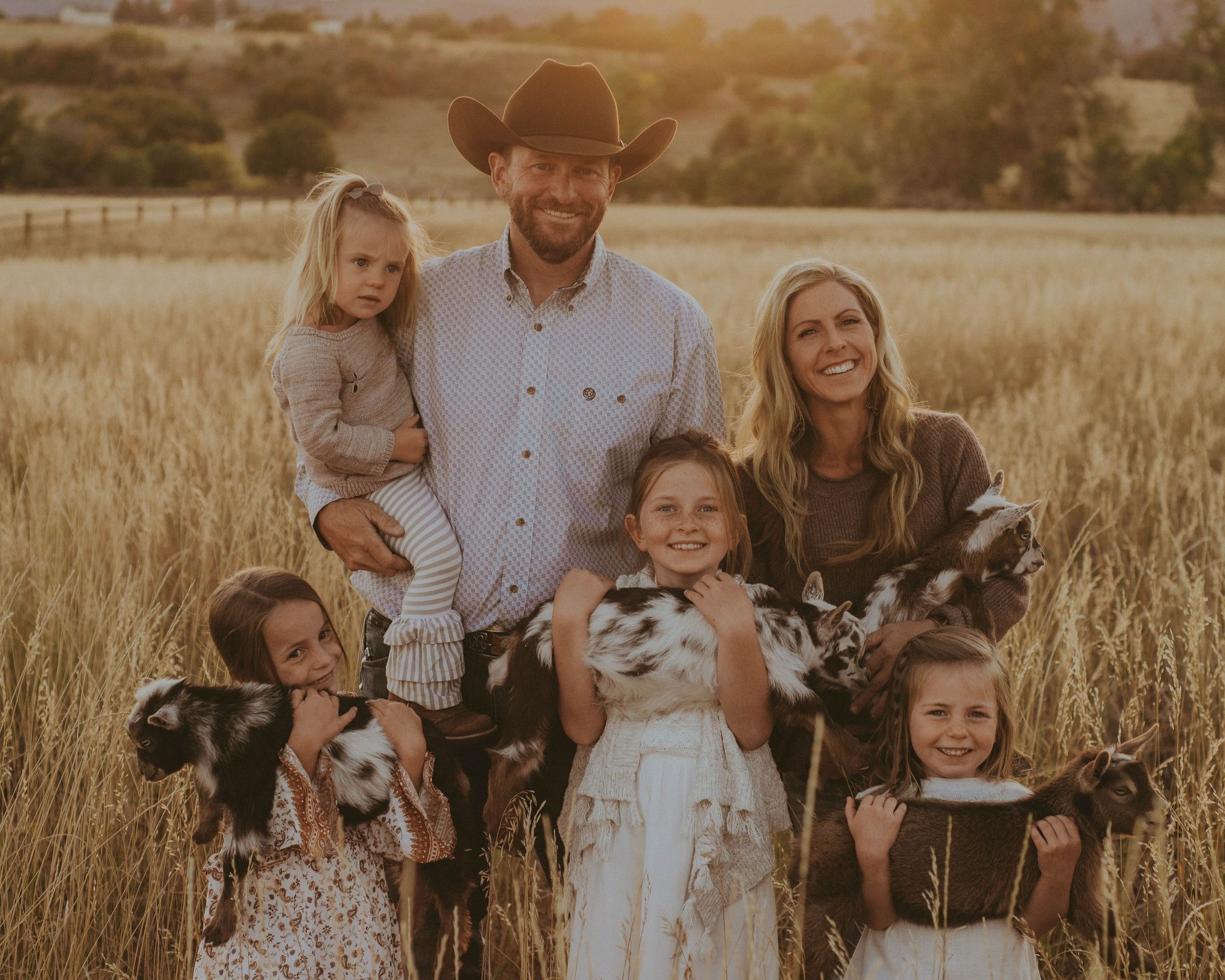 Family portrait in a golden field, featuring a smiling man in a cowboy hat holding a young girl, surrounded by four girls with goats, capturing a warm, joyful rural atmosphere.
